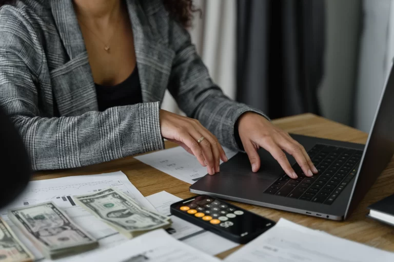 image of a woman's torso with a grey jacket, black top and a gold necklace on a laptop with papers, calulculator and money on the desk near her. Represents trying to reduce maintenance costs in her HOA