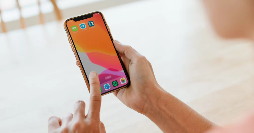 a hand holding a smartphone with an orange screensaver with 3 hoa communication apps: nextdoor, telegram and neigbrs by vinteum. It also has spotify, safari and image gallery, with a finger pointing at a screen