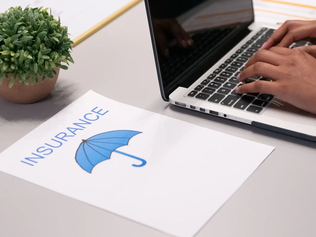 What Is Community Insurance? What You Don’t Know Can Hurt Your HOA 1 image of a grey desk with a little green plant, a laptop with hands resting on the keyboard and a page with a picture of a blue umbrella and the word insurance. Representing a HOA board member searching for community insurance