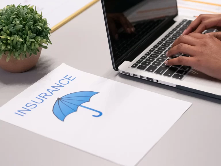 image of a grey desk with a little green plant, a laptop with hands resting on the keyboard and a page with a picture of a blue umbrella and the word insurance. Representing a HOA board member searching for community insurance