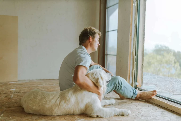 picture of a man with blonde hair looking out of a windom, with his arm around a fluffy white golden retriever