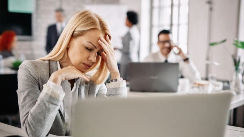 The image shows a woman in an office, with her hand in her forehead and a stressed look on her face due to Unethical HOA Board Members. 