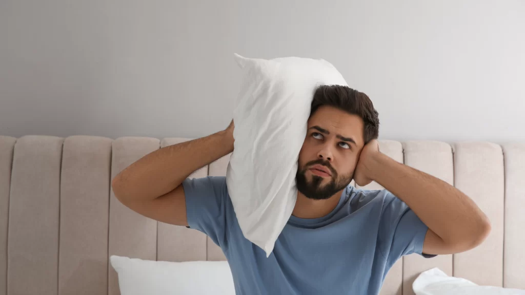 HOA Noise Complaints: 5 Steps to Handle Them Fairly and Fast 1 A frustrated man holds a pillow to his head, seeking comfort and contemplating a potential HOA noise complaints due to disruptive sounds in the background.