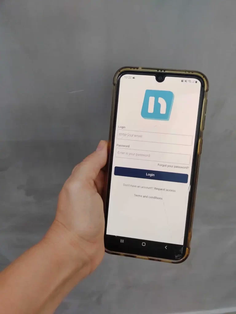 A white woman's hand holding an Android phone showing Neigbrs by Vinteum app login screen against a grey wall background