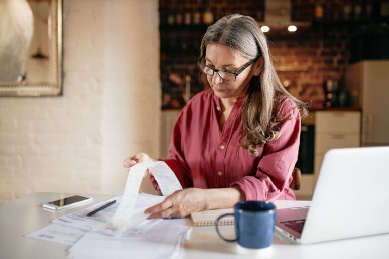 Serious focused middle aged female accountant in eyewear working from home providing outsource bookkeeping service, sitting at table with portable computer, calculating finances, doing paperwork